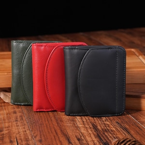 May include: A collection of three leather wallets in green, red, and black. Each wallet has a curved flap closure and is made of smooth leather. The wallets are arranged side by side on a wooden surface.