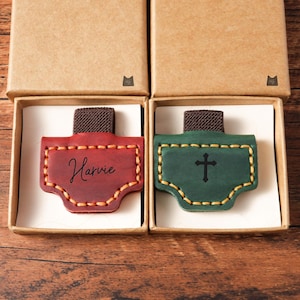 May include: Two leather bookmarks in boxes. One is red with the name "Harvie" in script, the other is green with a black cross. Both have brown elastic bands and are stitched with yellow thread.