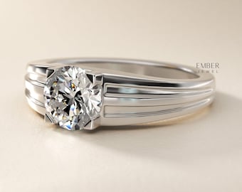May include: A silver ring featuring a large, round, clear gemstone. The ring has a simple, modern design with a grooved band. The gemstone is set in a square setting. The ring is displayed on a neutral background.