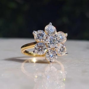 May include: A gold ring featuring a cluster of round, clear gemstones arranged in a flower shape. The ring's band is a polished gold color, and the gemstones sparkle brightly against a blurred background.