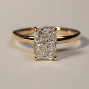 May include: A radiant-cut diamond ring with a gold band. The rectangular diamond is set with four prongs. The ring is set against a neutral background. The text "EMBER JEWEL" is visible on the ring.
