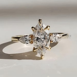 Peut inclure: Une bague de fiançailles en or avec une grosse pierre centrale de forme marquise et deux pierres latérales en forme de poire. La bague présente un anneau délicat et des griffes complexes. Le texte "EMBER JEWEL" est visible.