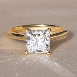 May include: A radiant-cut diamond engagement ring. The ring features a large, square-cut diamond set in a smooth, polished gold band. The diamond is held in place by four prongs. The ring is displayed on a neutral background.