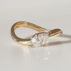 May include: A gold ring with a marquise-cut diamond. The ring has a unique, curved design. The diamond is set in a prong setting. The ring is on a white surface. The text "EMBER JEWEL" is visible on the ring.