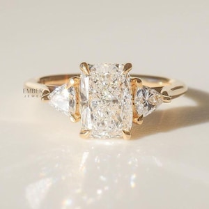 May include: A gold ring featuring a large, rectangular-cut diamond center stone, flanked by two smaller, triangular-cut diamonds. The ring has a delicate band and a classic, elegant design. The ring is set against a soft, neutral background.