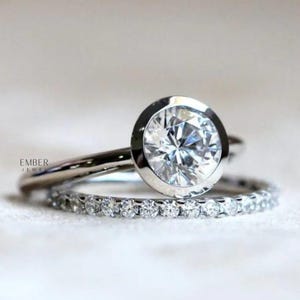 May include: Two silver rings. The top ring features a large, round, clear gemstone set in a bezel. The second ring is a thin band with small, clear gemstones. The rings are on a white background. The text "EMBER JEWEL" is visible.