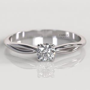 May include: A silver engagement ring with a clear, round diamond. The band has a twisted design. The ring is set against a white background. The ring is a classic style, suitable for an engagement.