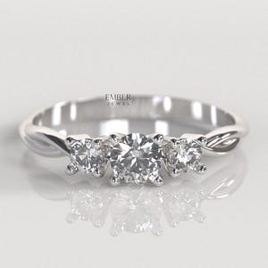 May include: A silver engagement ring with three clear, round-cut gemstones. The center stone is larger than the two side stones. The band has a twisted design. The text "EMBER JEWEL" is visible.