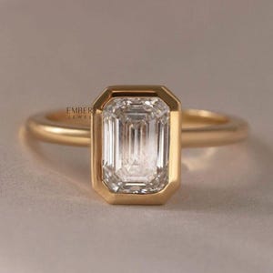 May include: A gold ring featuring an emerald-cut diamond. The diamond is set in a bezel setting, and the band is a simple, polished gold. The ring is displayed on a neutral background. The text "EMBER JEWEL" is visible.