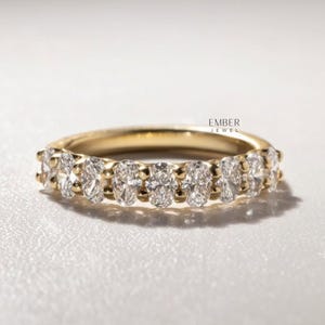 May include: A gold band ring featuring a row of oval-cut diamonds. The ring has a polished finish and is set against a soft, white background. The text "EMBER JEWEL" is visible.