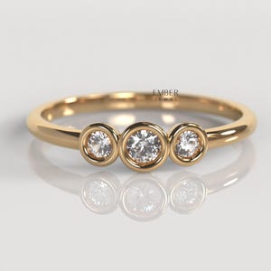 May include: A gold ring featuring three clear, round gemstones in bezel settings. The ring has a slender band and a polished finish. The gemstones are equally spaced across the top of the ring.