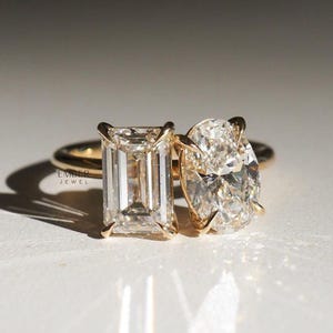 May include: Two gold rings with large, clear gemstones. One ring features an emerald-cut stone, the other an oval-cut stone. The rings are set on a white surface, with the text "EMBER JEWEL" visible.