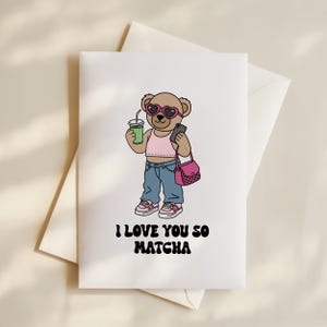 May include: A white greeting card featuring a cartoon bear in heart-shaped sunglasses, a pink top, and blue jeans. The bear holds a green drink and a phone, with a pink purse. The text "I LOVE YOU SO MATCHA" is below the bear.