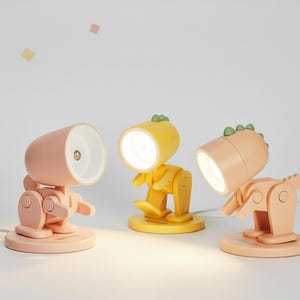 May include: Three whimsical desk lamps in the shape of dinosaurs. One is pink, one is yellow, and one is peach. Each lamp has a round head with a bright light and a body with articulated joints. The lamps are on a white background.
