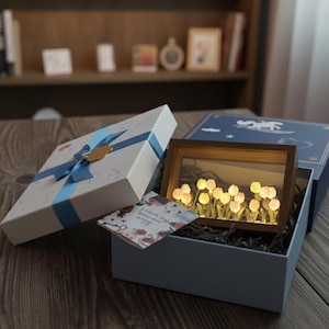 Könnte beinhalten: Eine dekorative Geschenkbox mit blauem Band und goldenem Anhänger. Im Inneren befindet sich eine Holz gerahmte, beleuchtete Tulpen-Anzeige in einer blauen Box. Eine kleine Karte mit Text ist ebenfalls enthalten.