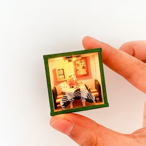 May include: Miniature diorama of a room with a table, chairs, and artwork, enclosed in a green-framed box. The scene is lit from within, creating a warm, inviting atmosphere. The diorama is held in a person's hand.