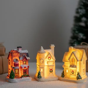 May include: Three illuminated miniature Christmas village houses. The houses are lit from within, showcasing details like windows, doors, and decorative elements. The houses are white with red, orange, and green accents. A Christmas tree is visible in the background.