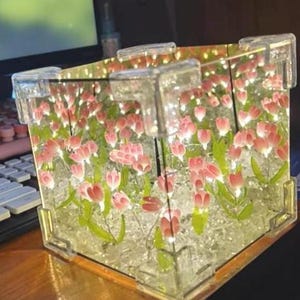 Puede incluir: Una caja decorativa cuadrada transparente con un diseño floral. La caja presenta flores rosas y blancas con forma de tulipán con tallos y hojas verdes. La caja está iluminada desde dentro, creando un brillo suave.