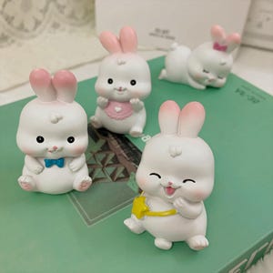 Puede incluir: Una colección de figuras de conejos blancos con orejas y detalles rosas. Cada conejo tiene una pose y expresión únicas, algunos con accesorios como una pajarita o un babero. Las figuras están sobre una superficie verde.