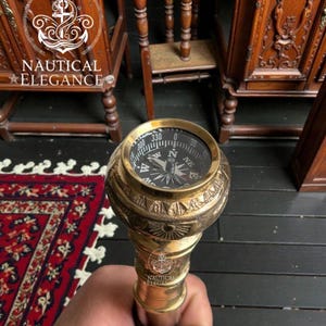 Puede incluir: Bastón de latón con brújula en la parte superior. La brújula tiene una esfera negra con marcas blancas y una aguja que apunta al norte. El bastón se sostiene en la mano de una persona. El texto "NAUTICAL ELEGANCE" es visible.