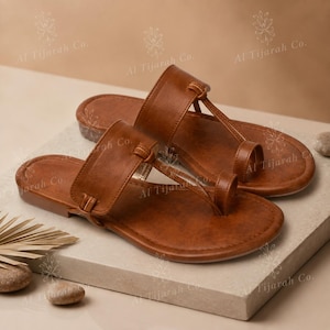 May include: A pair of brown flat sandals featuring a wide strap across the midfoot and a toe loop design. The sandals have a smooth, solid-colored finish with a rounded toe bed and a flat sole. The text Al Tijarah Co. is visible on the image.