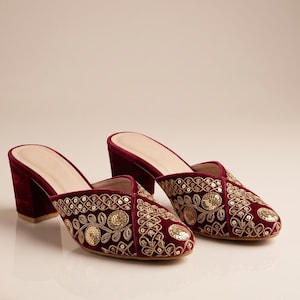 May include: A pair of deep burgundy velvet mules featuring a block heel and a pointed toe. The upper surface is adorned with intricate gold embroidery and circular gold sequins arranged in a decorative floral and geometric pattern throughout.