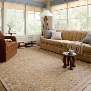 May include: A cozy living room with a tan patterned rug, a brown leather armchair, and a tan sofa with throw pillows. A wooden side table holds a teacup. Large windows with blue and tan shades provide natural light.