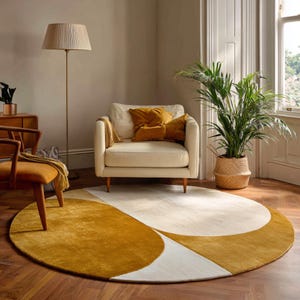 May include: A circular rug with a geometric pattern in mustard yellow, cream, and white. The rug is on a wooden floor in a living room. A cream armchair with mustard cushions is nearby. A floor lamp and a potted plant are also visible in the room.