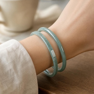 Pode incluir: Dois braceletes de jade verde-pálido, lisos e usados juntos. Um dos braceletes apresenta uma pequena incrustação metálica retangular de tom prateado, inserida nivelada na superfície de pedra polida. Os braceletes têm um perfil arredondado.