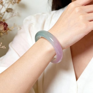 May include: A translucent bangle bracelet with a gradient of colors, transitioning from a soft green to a light purple. The smooth, polished surface reflects light, highlighting the bracelet's elegant design. The bracelet is worn on a person's arm.