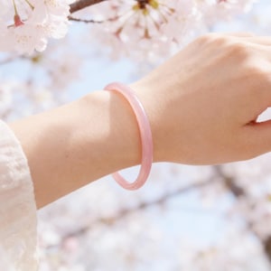 May include: A light pink bangle bracelet. The bracelet is a solid, circular band. The image shows the bracelet on a person's arm, with a blurred background of white and pink flowers.