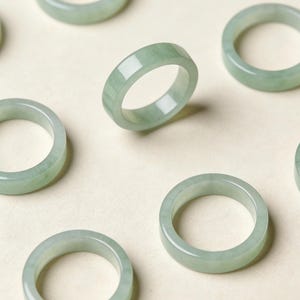 Peut inclure: Plusieurs bagues en jade vert clair sont présentées sur un fond neutre. Les bagues ont une surface lisse et polie et un design simple et circulaire. Les bagues sont de couleur vert pâle.