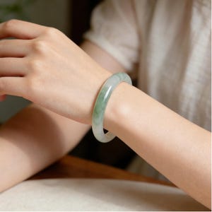 Puede incluir: Un brazalete de jade con una superficie lisa y pulida. El brazalete presenta una mezcla de tonos verde pálido y blanco, creando un efecto veteado. La joya se lleva en la muñeca.