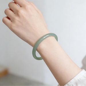 May include: A light green jade bangle bracelet. The smooth, polished bracelet is circular and worn on a wrist. The jade has a translucent quality, with subtle variations in color. The background is a soft, neutral tone.