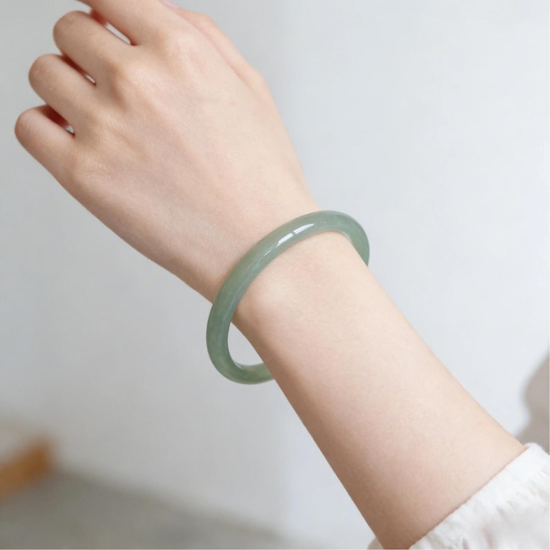 May include: A light green jade bangle bracelet. The smooth, polished bracelet is circular and worn on a wrist. The jade has a translucent quality, with subtle variations in color. The background is a soft, neutral tone.