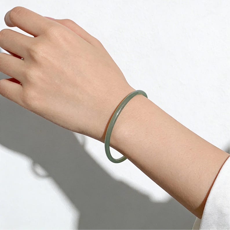 May include: A light green jade bangle bracelet. The bracelet is a solid, smooth band of translucent jade, showcasing a subtle, natural color variation. The bracelet is worn on a wrist.