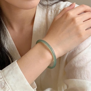 May include: A light green jade bangle bracelet. The smooth, polished bracelet is a pale green color and is worn on the wrist. The bracelet is a simple, elegant piece of jewelry.