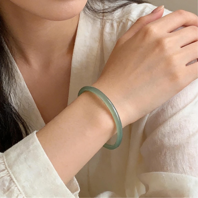 May include: A light green jade bangle bracelet. The smooth, polished bracelet is a pale green color and is worn on the wrist. The bracelet is a simple, elegant piece of jewelry.