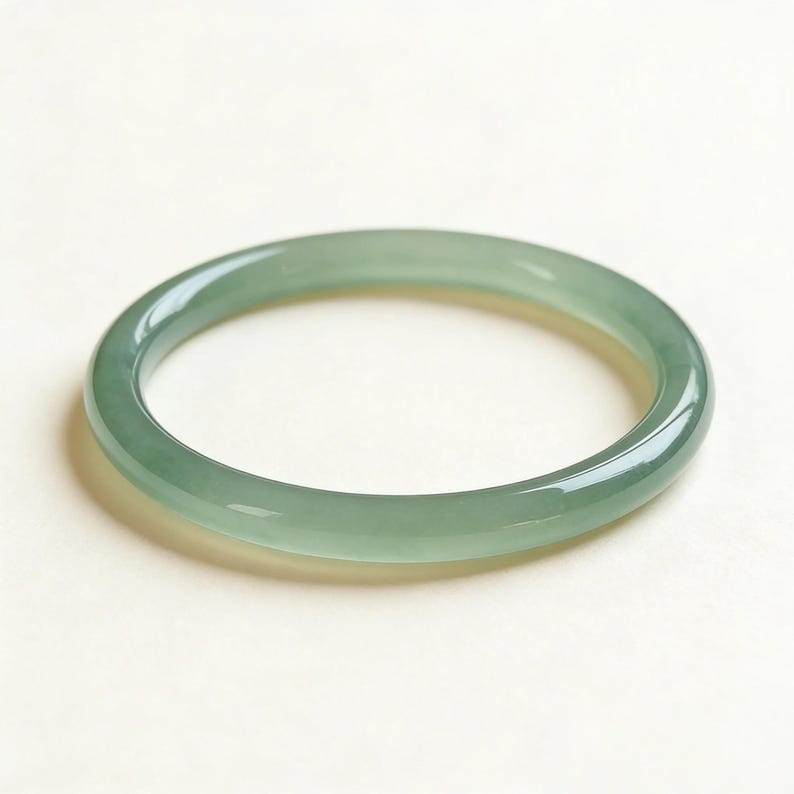 May include: A smooth, jade green bangle bracelet. The circular bracelet is a solid color with a glossy finish. The jade is a light green color. The bracelet is a simple, elegant piece of jewelry.