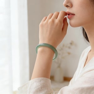 May include: A light green jade bangle bracelet. The smooth, polished bracelet is worn on a wrist. The jade has a translucent quality, and the color is a soft, muted green. The bracelet is a classic piece of jewelry.