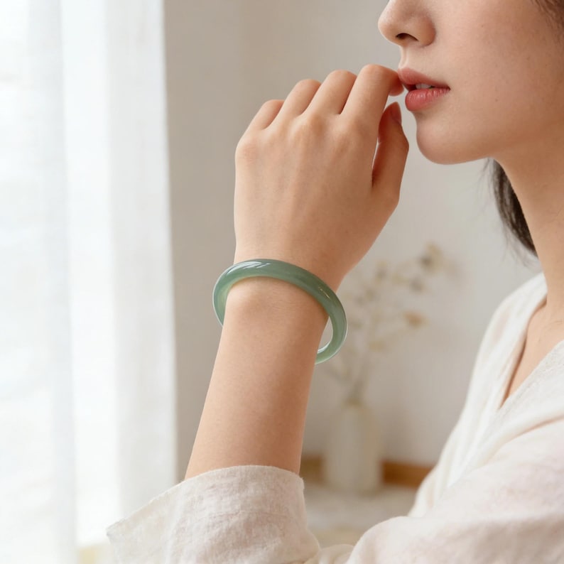 May include: A light green jade bangle bracelet. The smooth, polished bracelet is worn on a wrist. The jade has a translucent quality, and the color is a soft, muted green. The bracelet is a classic piece of jewelry.