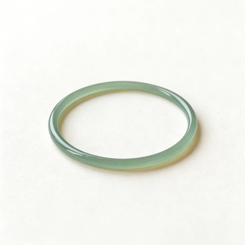 May include: A light green jade bangle bracelet. The smooth, circular bracelet is a pale green color and appears to be made of a translucent stone. The bangle is displayed on a white background.