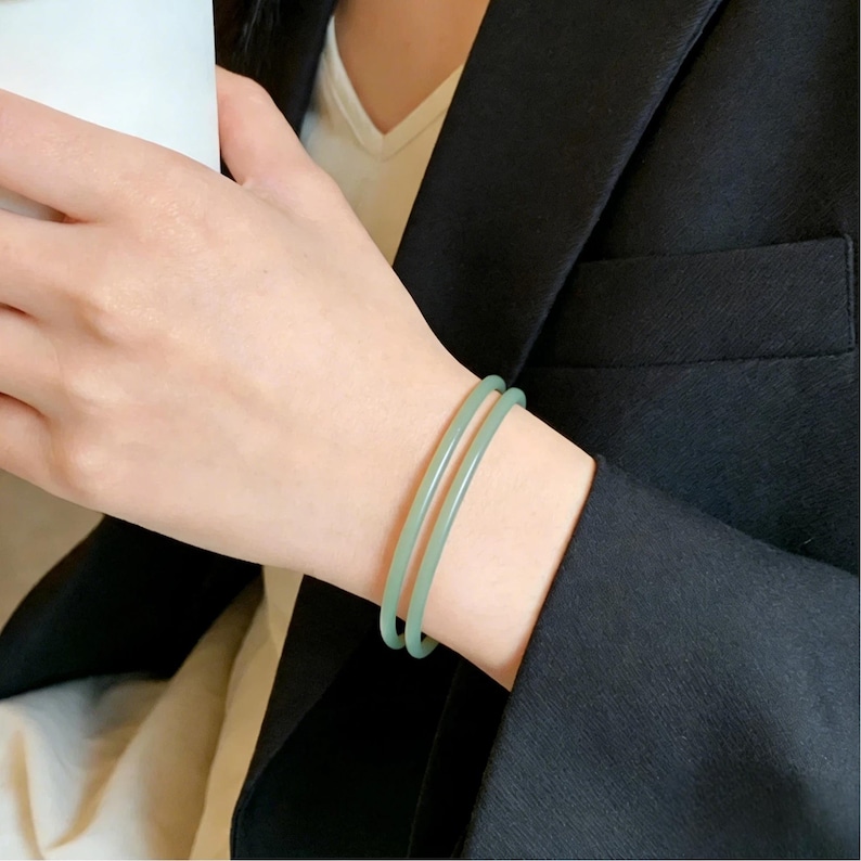 May include: Two light green jade bangles are worn on a wrist. The smooth, round bracelets are a soft, muted green color. The person is wearing a black blazer and a white shirt.