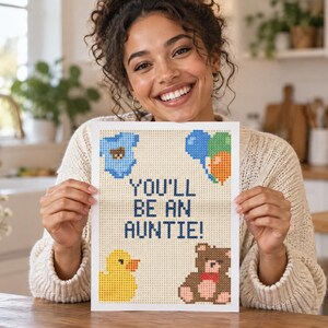 May include: A rectangular print featuring a pixelated design on a light background with the text YOU'LL BE AN AUNTIE! in dark blue. The design includes a blue baby onesie, a cluster of blue, green, and orange balloons, a yellow duck, and a brown teddy bear.