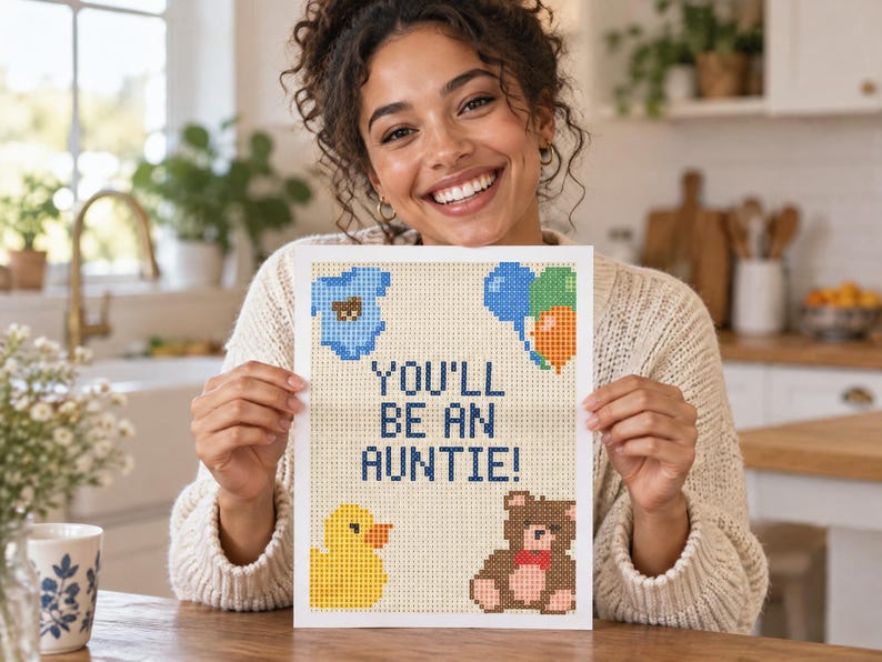 May include: A rectangular print featuring a pixelated design on a light background with the text YOU'LL BE AN AUNTIE! in dark blue. The design includes a blue baby onesie, a cluster of blue, green, and orange balloons, a yellow duck, and a brown teddy bear.