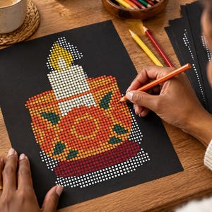 May include: A black sheet of paper featuring a pixel art design of a lit candle in a decorative holder. The candle has a yellow flame and white wax, while the holder is orange with dark green leaf accents and a deep red base.