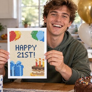 May include: A rectangular diamond painting card featuring a pixelated design with the text HAPPY 21ST! in blue. The card displays colorful balloons, a blue gift box, and a birthday cake with lit candles against a light tan grid background.