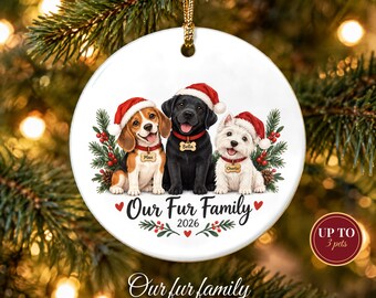 May include: A round white ceramic Christmas ornament featuring a beagle, a black Labrador, and a white terrier wearing red Santa hats. Each dog wears a red collar with a bone-shaped tag labeled Max, Bella, or Charlie. Text reads Our Fur Family 2026.