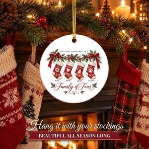 May include: A round white ceramic Christmas ornament featuring a graphic of four red and white stockings hanging from a green garland with red bows. Text reads: Our First Christmas as a Family of Four, Dad, Mom, Emma, Buddy, and 2026.
