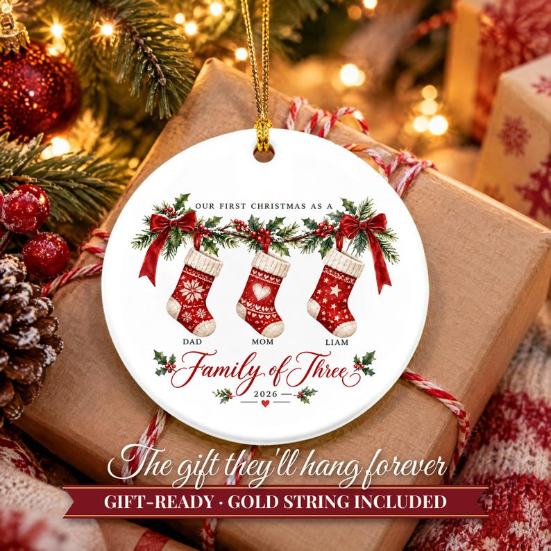 May include: A round white ceramic ornament featuring a festive illustration of three red Christmas stockings hanging from a holly garland with red bows. The text reads Our First Christmas as a Family of Three 2026 with Dad, Mom, and Liam labels.